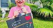 Noah Thompson is registered to the contest to win money with this photo: child, colorful, daylight, first_day, grass, greenery, happy, holding, outdoor, palm_tree, person, portrait, pre_k, school, shirt, sign, smiling, sunny, writing, young