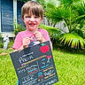 child, colorful, daylight, first_day, grass, greenery, happy, holding, outdoor, palm_tree, person, portrait, pre_k, school, shirt, sign, smiling, sunny, writing, young