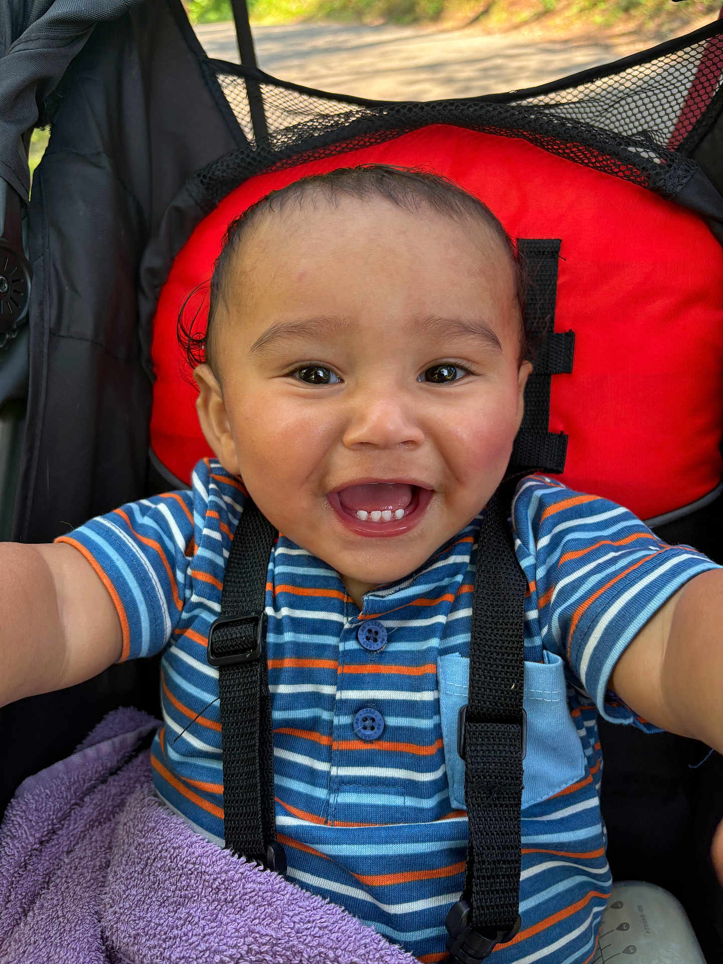 Zaiden is registered to the contest to win money with this photo: baby, child, smiling, striped_shirt, blue_shirt, orange_stripes, stroller, red_backrest, purple_towel, happy, face, teeth, person, infant, outdoor, daylight, closeup, cute, baby_boy, seatbelt