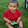 toddler, child, grass, red_shirt, toy, ladybug_toy, outdoor, sitting, curious_expression, short_hair, black_socks, green_background, cartoon_prints, baby, playtime, cute, natural_light, person, summer, casual_clothing