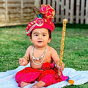 Kriday joined the competition — help win amazing prizes! baby, child, traditional_clothing, red_turban, peacock_feather, beads, necklace, bracelet, staff, grass, blanket, outdoor, cute, portrait, sitting, festive, earrings, happy, decorative, young