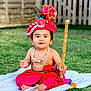 baby, child, traditional_clothing, red_turban, peacock_feather, beads, necklace, bracelet, staff, grass, blanket, outdoor, cute, portrait, sitting, festive, earrings, happy, decorative, young