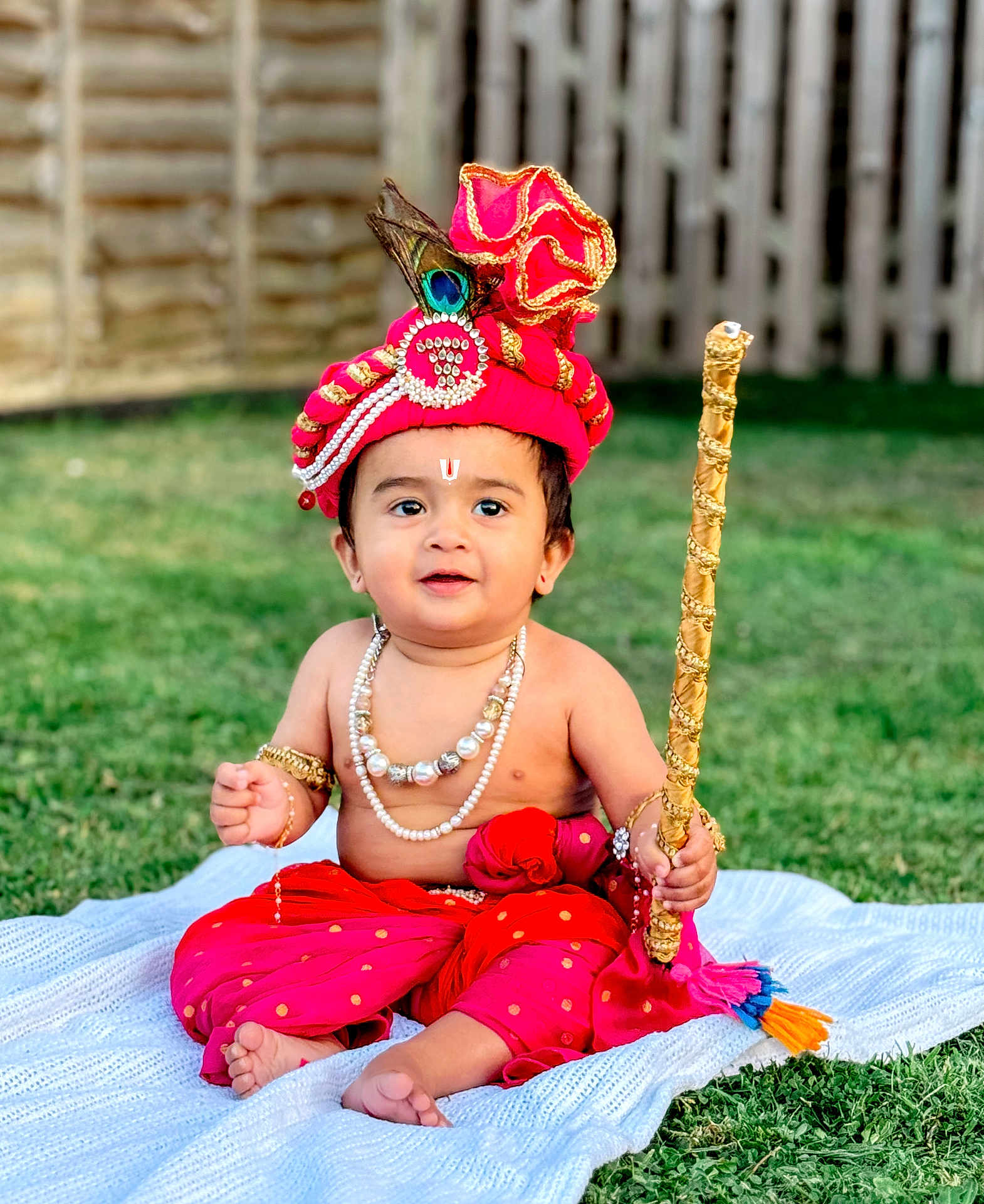 Kriday joined the competition — help win amazing prizes! baby, child, traditional_clothing, red_turban, peacock_feather, beads, necklace, bracelet, staff, grass, blanket, outdoor, cute, portrait, sitting, festive, earrings, happy, decorative, young