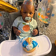 Saage is registered to the contest to win money with this photo: baby, child, party_hat, highchair, cupcake, frosting, pacifier, birthday, celebration, balloons, confetti, smile, clothing, face, hand, table, indoor, decorations, food, happy