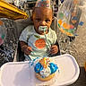 baby, child, party_hat, highchair, cupcake, frosting, pacifier, birthday, celebration, balloons, confetti, smile, clothing, face, hand, table, indoor, decorations, food, happy