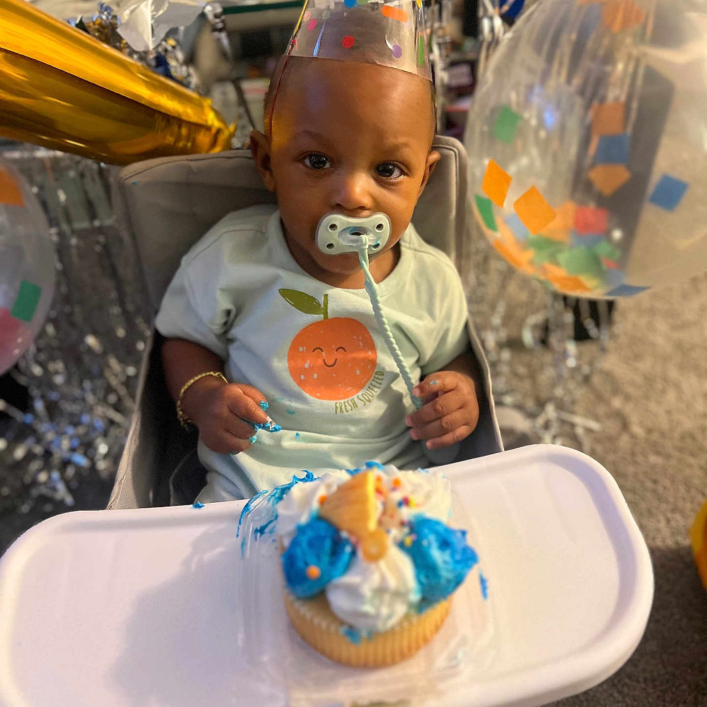 Saage is registered to the contest to win money with this photo: baby, child, party_hat, highchair, cupcake, frosting, pacifier, birthday, celebration, balloons, confetti, smile, clothing, face, hand, table, indoor, decorations, food, happy