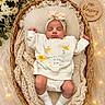 baby, newborn, sleeping, basket, blanket, flower, headband, fairy_lights, wooden_plaque, cozy, soft_texture, white_clothing, knitted, decor, peaceful, infant, cute, indoors, portrait, resting
