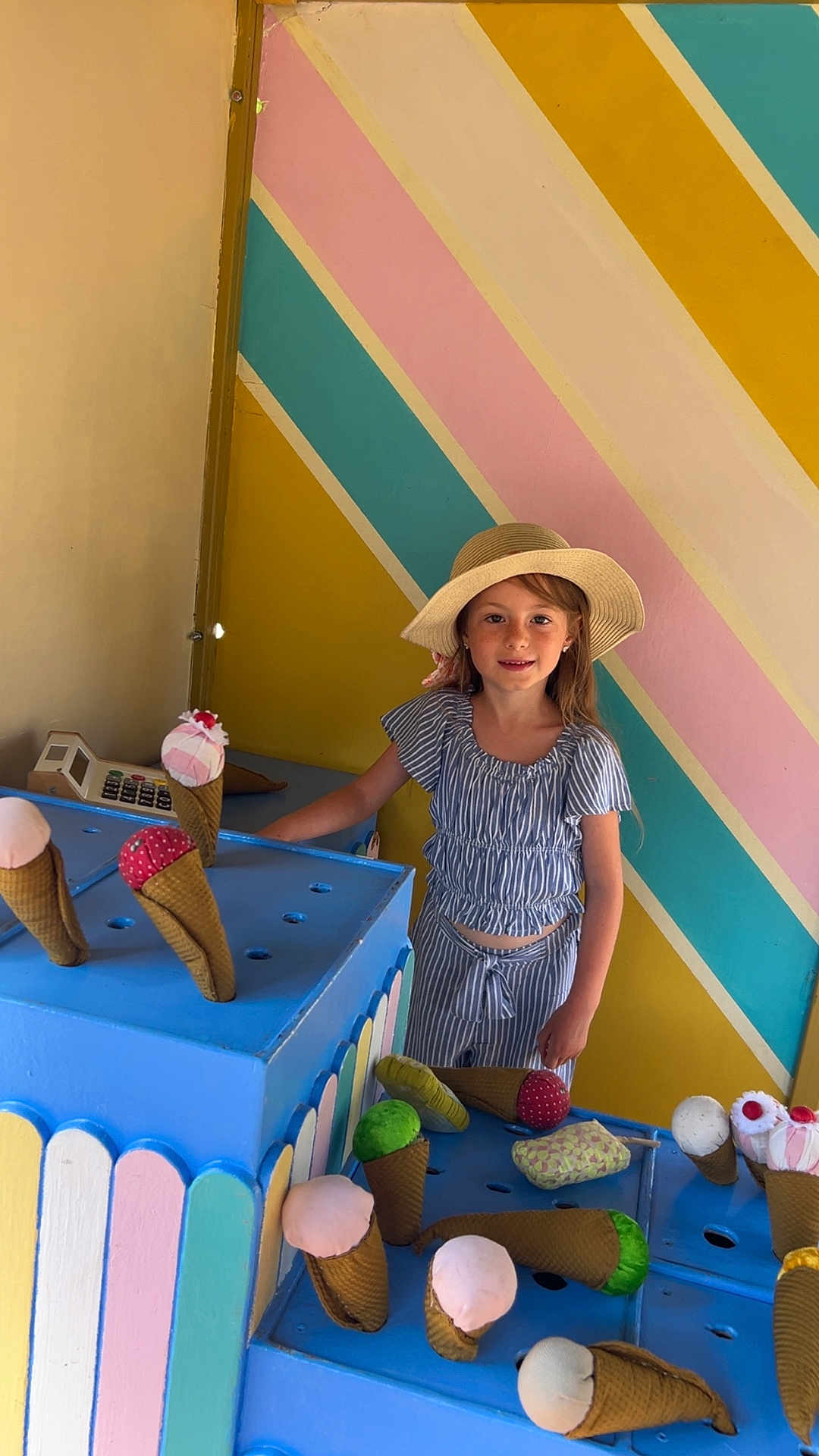Maëlys a rejoint le concours — aidez-le/la à gagner de superbes lots ! child, girl, striped_outfit, sunhat, toy_ice_cream, play_area, blue_shelves, colorful_wall, indoor, smiling, pretend_play, fabric_toys, ice_cream_cones, playtime, happy, cute, summer, casual_clothing, toy_cash_register, playful