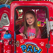 Maëlys a rejoint le concours — aidez-le/la à gagner de superbes lots ! child, girl, amusement_ride, paw_patrol, red_vehicle, smiling, long_hair, pink_shirt, carnival, fairground, playful, colorful, metal, seat, window, door, handle, fun, outdoor, daytime