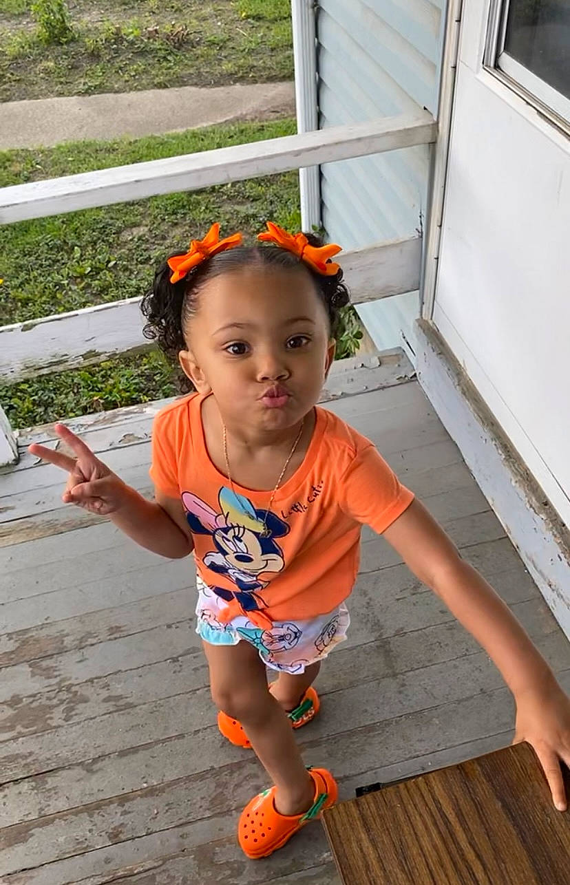 Kalee is registered to the contest to win money with this photo: baby_toddler_clothing, child, eye, fence, flooring, fun, grass, happy, hardwood, head, human_leg, leisure, orange, person, plant, sitting, skin, t_shirt, thigh, toddler