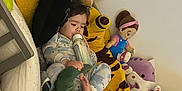 Lilith is registered to the contest to win money with this photo: child, baby, pajamas, bottle, stuffed_animals, plush_toys, bed, giraffe, dinosaur, doll, sleepwear, toy, comfort, indoor, cute, resting, soft, colorful, cozy, young