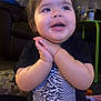 toddler, child, smiling, hands_clasped, indoor, black_tshirt, patterned_shirt, curly_hair, couch, pillow, carpet, person, happy, cute, baby, sitting, furniture, face, expression, playful
