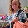 accessory, bag, bed, blonde_hair, casual, child, clothing, clutter, curly_hair, fabric, girl, hand, indoor, person, portrait, room, sitting_area, smile, textile, wall