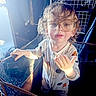 toddler, child, glasses, playpen, indoor, curly_hair, onesie, light, hand, face, person, expression, home, furniture, baby, clothing, portrait, standing, cute, curiosity