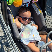 Gabriel joined the competition — help win amazing prizes! baby, child, infant, sunglasses, shopping_cart, grocery_store, outdoor, sunlight, toys, baby_bottle, blanket, seated, portrait, smiling, vehicle, metal, cart, parking_lot, casual, family