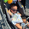 baby, child, infant, sunglasses, shopping_cart, grocery_store, outdoor, sunlight, toys, baby_bottle, blanket, seated, portrait, smiling, vehicle, metal, cart, parking_lot, casual, family