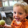child, toddler, smiling, indoor, orange_shirt, dinosaur_print, ball, toy, blond_hair, happy, carpet, sofa, window, curtains, wood_floor, living_room, person, face, cute, playful