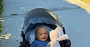 Noah is registered to the contest to win money with this photo: baby, child, infant, stroller, smiling, outdoor, sidewalk, park, grass, sunlight, plush_toy, bunny, toy, blue_clothing, portrait, sitting, happy, face, wheels, fence