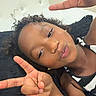 Taisha is registered to the contest to win money with this photo: child, portrait, curly_hair, peace_sign, smiling, face, eyes, lips, indoor, bed, pillow, bedroom, black_clothing, striped_clothing, sitting, lying_down, close_up, selfie, hand_gesture, relaxed