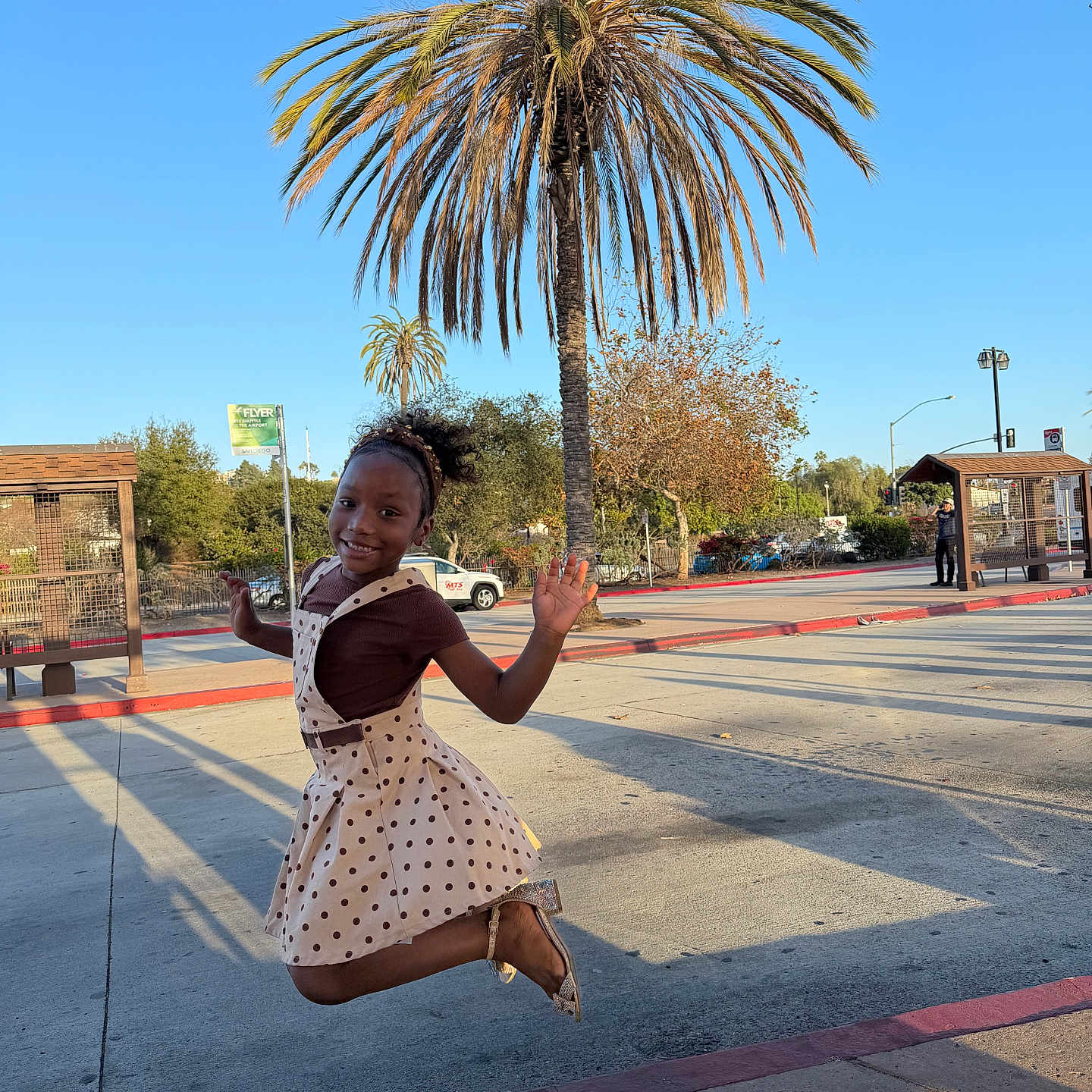 Taisha is registered to the contest to win money with this photo: child, girl, smiling, jumping, outdoor, palm_tree, street, sidewalk, bus_stop, urban, sunny, blue_sky, dress, polka_dot, waving, portrait, playful, joyful, shadow, photography