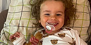 Kaïs participe au concours pour gagner de l'argent avec cette photo : bed, blanket, child, clothing, cozy, curly_hair, cute, face, happy, indoor, lying_down, pacifier, pajamas, person, pillow, smiling, teddy_bear, toddler, toy, young_child