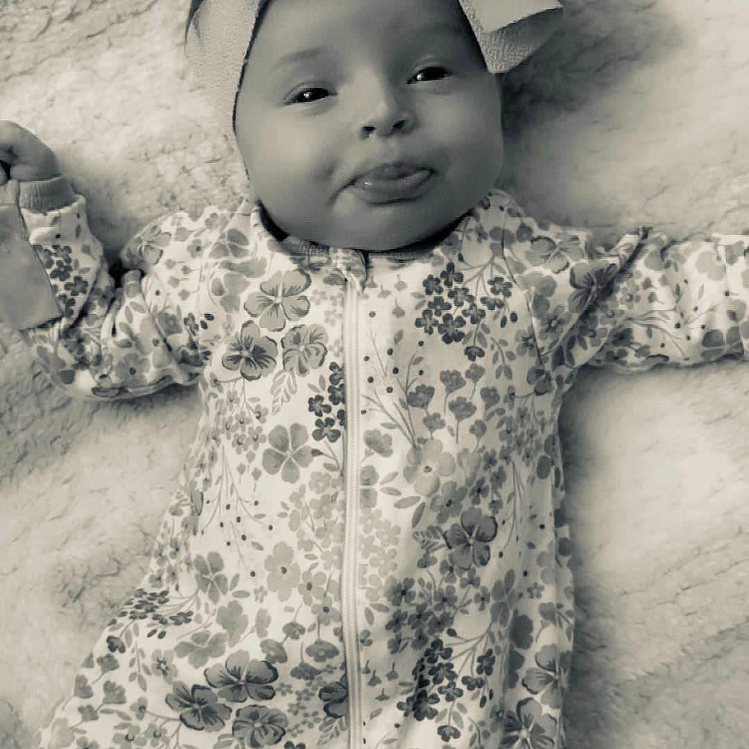 Renesemae Raiyne is registered to the contest to win money with this photo: baby, blanket, bow, child, comfortable, cute, face, floral_pattern, happy, headband, indoors, infant, lying_down, onesie, playful, portrait, smiling, soft_texture, tongue_out, young