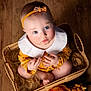 baby, infant, child, basket, headband, yellow_clothing, white_collar, blue_eyes, wooden_floor, flowers, orange_flowers, woven_basket, soft_fibers, cute, portrait, sitting, looking_up, indoor, adorable, hands
