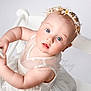 Rosanna is registered to the contest to win money with this photo: baby, child, blue_eyes, flower_crown, white_dress, chair, cute, portrait, infant, hands, looking_up, studio, soft_lighting, curious, skin, headband, young, person, indoors, fair_skin