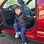 child, boy, car, door, sneakers, clothing, black_vest, blue_pants, outdoor, asphalt, vehicle_interior, seat, curious_expression, casual, daylight, holding_door, young_child, small_person, stepping_out, red_car
