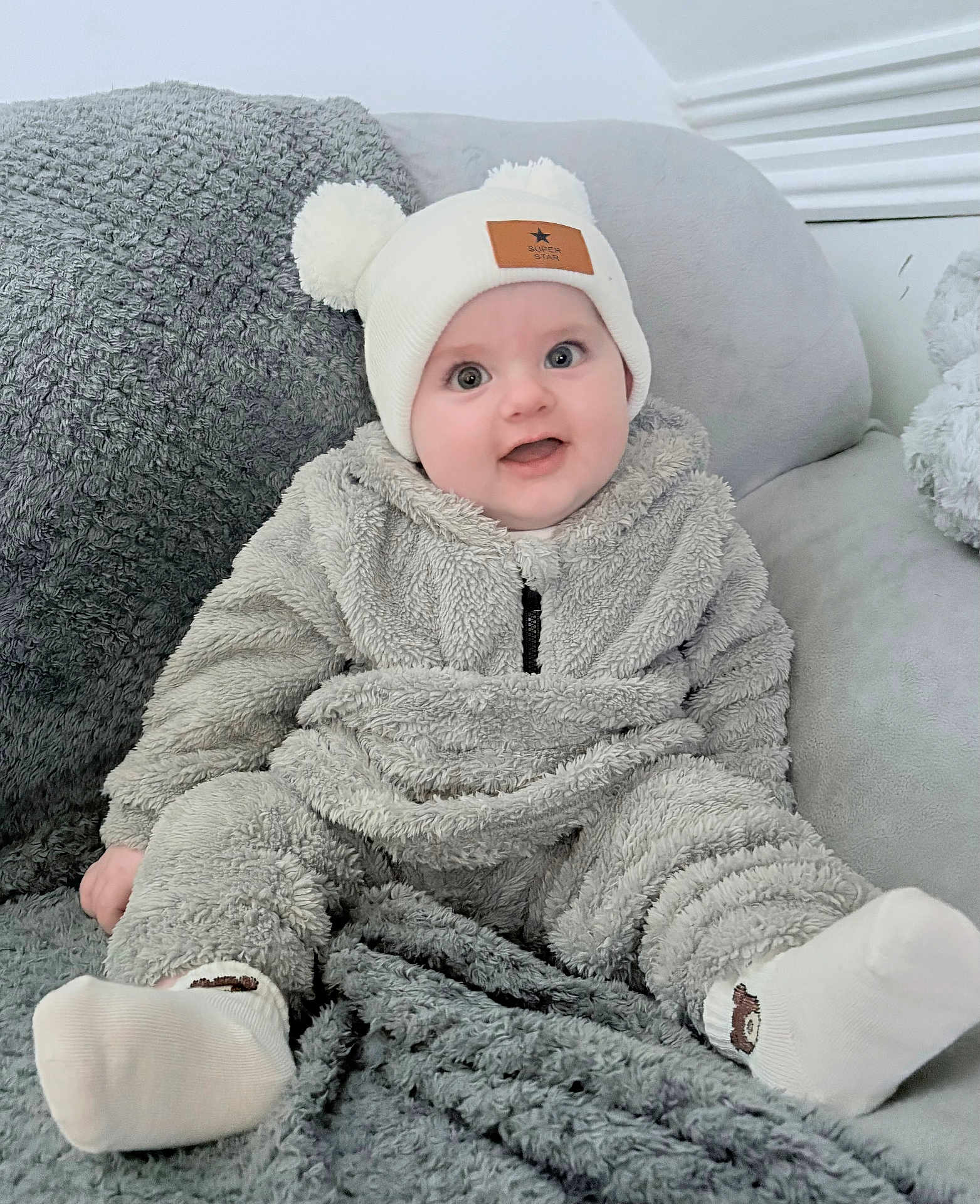 Enzo joined the competition — help win amazing prizes! baby, infant, child, smiling, hat, pom_poms, fluffy_clothing, socks, couch, blanket, soft_texture, indoor, comfortable, cute, happy, portrait, seated, cozy, warm_clothing, headwear