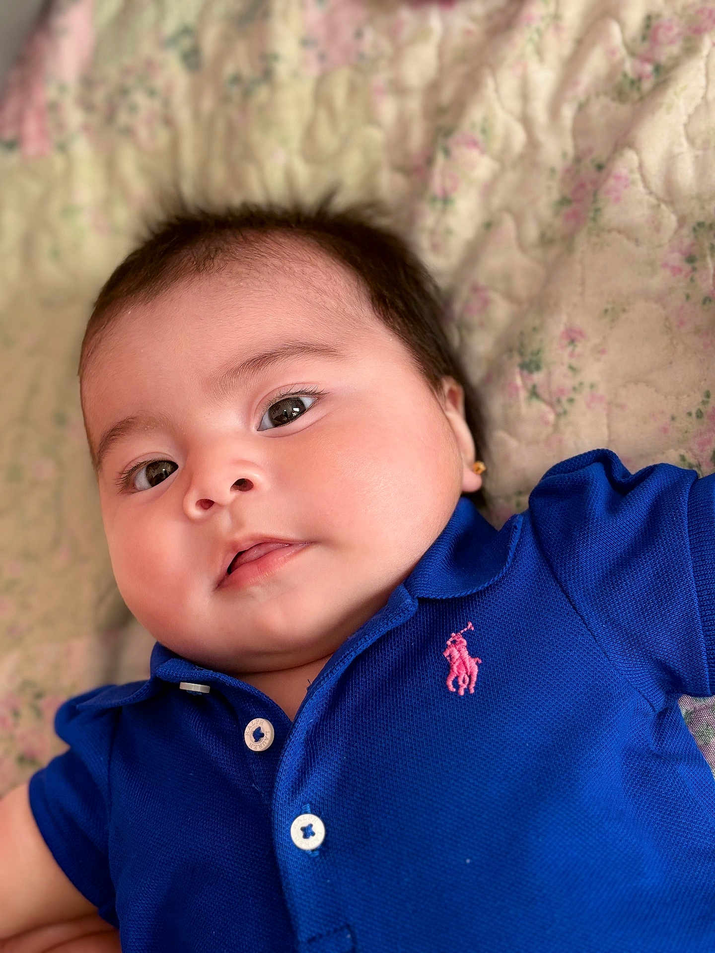 Cattleya is registered to the contest to win money with this photo: baby, child, face, eyes, polo_shirt, blue_clothing, quilt, floral_pattern, infant, portrait, cute, soft_lighting, closeup, skin, head, earrings, young_child, indoor, lying_down, adorable