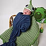 newborn, baby, sleeping, knitted_hat, blue_cloth, green_blanket, dinosaur_blanket, crocheted_toy, green_toy, baby_wrapped, infant, cute, studio_photo, white_background, peaceful, soft_texture, child, portrait, cozy, handmade