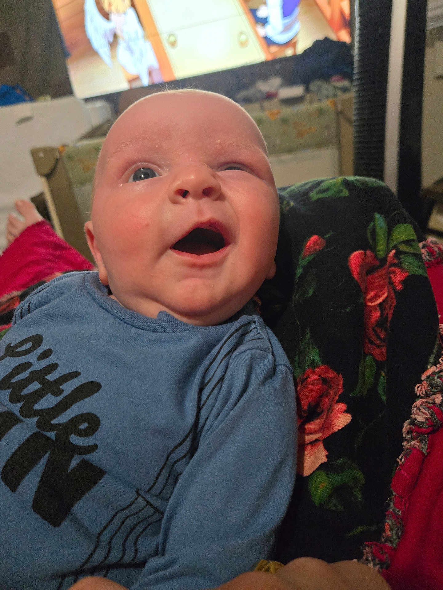 Wylder is registered to the contest to win money with this photo: baby, infant, child, portrait, mouth_open, surprised, blue_shirt, blanket, floral_blanket, television, living_room, sofa, face, head, eyes, newborn, skin, clothing, home, candid
