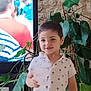 background_blur, casual_clothing, child, decor, face, furniture, greenery, happy, home, indoor, light, living_room, palm_tree_pattern, person, plant, short_hair, smiling, television, white_shirt, young_child