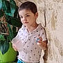 blue_pants, boy, casual_clothing, child, cup, curious, greenery, home, indoor, leisure, natural_light, plant, portrait, potted_plant, short_hair, standing, stone_wall, toy, white_shirt, young_child