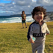 Iziah is registered to the contest to win money with this photo: active, casual_clothing, child, cloudy_sky, coast, daytime, fun, grass, happy, jordan, nature, ocean, outdoor, pacifier, playing, running, smiling, toddler, tracksuit, young_child