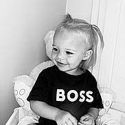 Kahïna participe au concours pour gagner de l'argent avec cette photo : bed, black_and_white, casual_clothing, child, cute, expression, furniture, hair_tied, indoor, person, pillow, plush_toy, portrait, sitting, small_object, smiling, text_on_clothing, toddler, tshirt, young_child