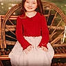 child, girl, smiling, portrait, sitting, wicker_chair, red_sweater, white_skirt, long_hair, indoors, vintage, warm_lighting, soft_focus, furniture, hands, dress, face, person, nostalgic, photograph