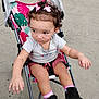 toddler, child, stroller, curly_hair, pink_bows, gray_shirt, pink_shorts, pink_socks, black_shoes, outdoor, concrete, curious, sitting, baby, person, footwear, clothing, cute, infant, baby_carriage