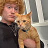 Sativa joined the competition — help win amazing prizes! person, cat, indoor, curly_hair, ginger_cat, collar, bell, holding, face, pet, animal, young_adult, expression, wall, window, black_shirt, necklace, red_hair, fur, home