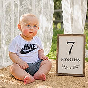 Messiah is registered to the contest to win money with this photo: baby, infant, child, blue_eyes, nike_tshirt, shorts, sitting, outdoor, grass, sign, 7_months, lace_curtain, wooden_frame, mat, cute, portrait, sunlight, natural_light, expression, young