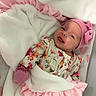 baby, baby_clothing, bedding, blanket, bow, closeup, cozy, crib, eyes_open, floral_outfit, indoors, infant, mittens, newborn, pink_hat, portrait, smiling, soft_lighting, swaddle, teddy_bear