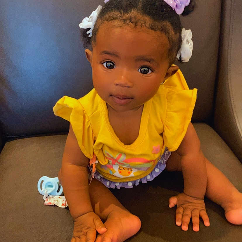 Emerie is registered to the contest to win money with this photo: toddler, child, baby, yellow_clothing, purple_hair_bows, brown_couch, indoor, cute, portrait, sitting, small_hands, barefoot, looking_at_camera, innocent_expression, hair_accessories, young_child, bright_eyes, casual, home, furniture