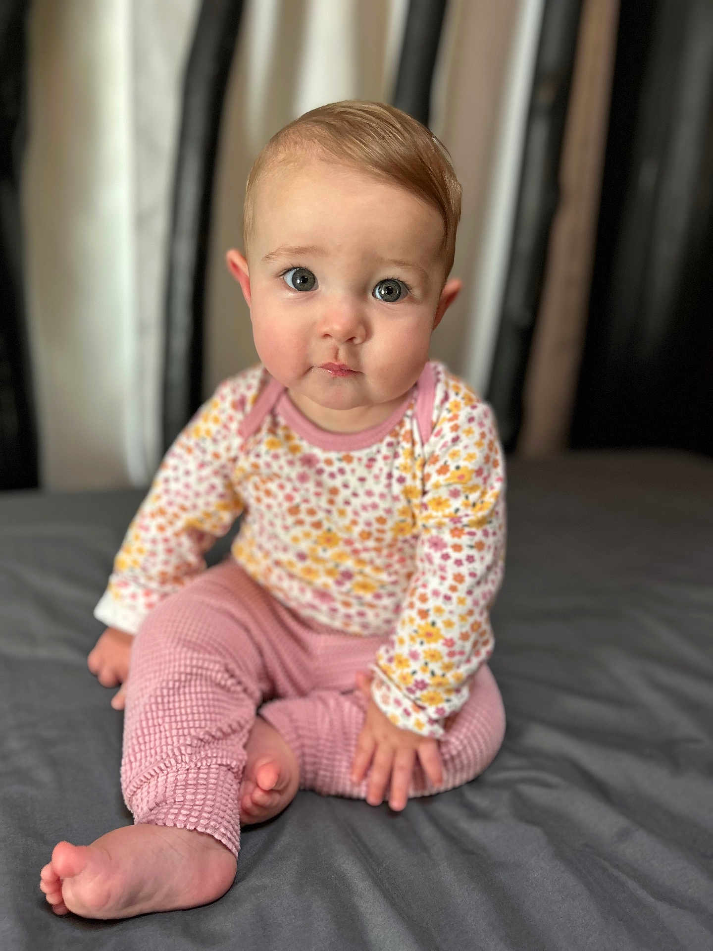 Britton is registered to the contest to win money with this photo: baby, child, infant, sitting, barefoot, pink_pants, floral_shirt, curly_hair, large_eyes, indoor, portrait, soft_light, bed, gray_bedspread, cute, looking_at_camera, young_child, close_up, skin, human