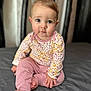 baby, child, infant, sitting, barefoot, pink_pants, floral_shirt, curly_hair, large_eyes, indoor, portrait, soft_light, bed, gray_bedspread, cute, looking_at_camera, young_child, close_up, skin, human