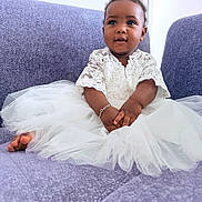 Jalia a rejoint le concours — aidez-le/la à gagner de superbes lots ! baby, child, white_dress, lace, bracelet, purple_couch, sitting, curly_hair, indoor, portrait, smiling, cute, infant, hands, feet, soft_texture, cozy, young, person, tulle