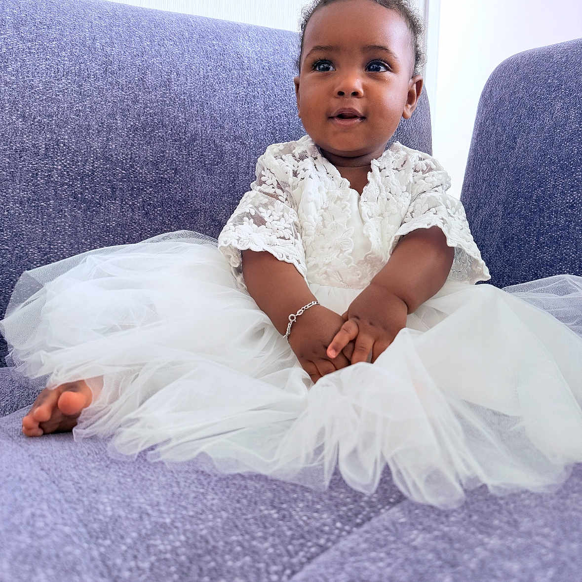 Jalia a rejoint le concours — aidez-le/la à gagner de superbes lots ! baby, bracelet, child, cozy, curly_hair, cute, feet, hands, indoor, infant, lace, person, portrait, purple_couch, sitting, smiling, soft_texture, tulle, white_dress, young