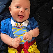 Mckenzie is registered to the contest to win money with this photo: baby, blanket, blue_clothing, child, closeup, costume, cute, face, happy, human, indoor, infant, lying_down, playful, portrait, red_bow, smiling, tongue_out, yellow_skirt, young
