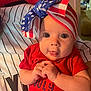 american_flag, baby, bow, child, closeup, clothing, cute, eyes, face, hand, headwrap, holding, indoor, infant, patriotic, person, portrait, red_shirt, striped_jersey, tongue_out