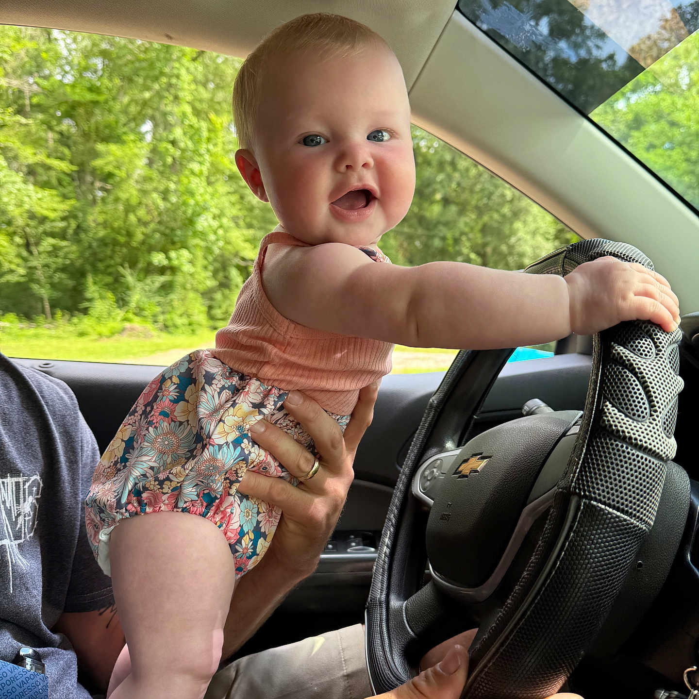 Opal is registered to the contest to win money with this photo: adult_hand, baby, car, cheerful, child, cute, driving, floral_clothing, greenery, happy, infant, interior, outdoor, person, portrait, seat, smiling, steering_wheel, summer, window